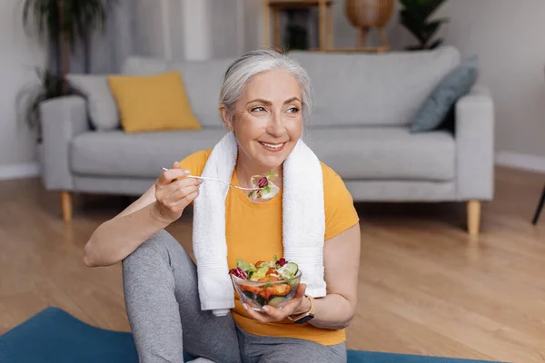 Friska livsstil koncept. Glad senior kvinna äter färsk grönsakssallad, sitter på yogamatta efter hemmaträning — Stockfoto