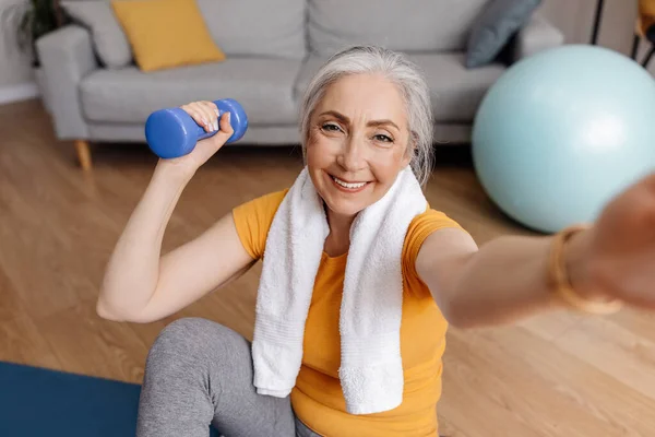 Emocionada mujer mayor tomando selfie mientras hace ejercicio con pesas en casa, sentada en una esterilla de yoga — Foto de Stock