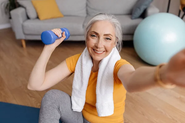 Evde dambıllarla egzersiz yaparken, yoga minderinde otururken selfie çeken heyecanlı son sınıf kadını. — Stok fotoğraf