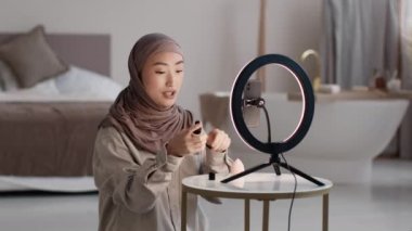 Genç Müslüman Asyalı kadın tesettürlü ve tesettürlü. Güzellik blogu için video çekiyor. Evde telefon kamerasında, yeni bir ton kremi reklamı yapıyor.
