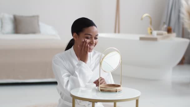 美容美发概念 身穿浴衣的年轻而快乐的非洲女人在脸上涂上水疗霜 对着镜子微笑 坐在浴室里 慢动作 — 图库视频影像