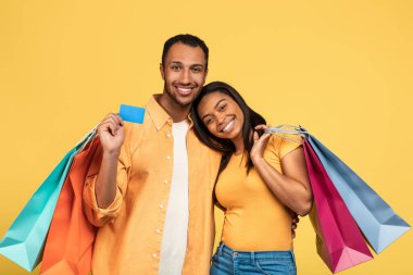 Alışveriş, perakende, satış ve indirim konsepti. Bir sürü parlak alışveriş çantası ve kredi kartı tutan mutlu genç siyah çift sarı stüdyo arka planında elektronik para kullanıyorlar.