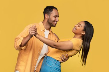 Sarı stüdyo arka planında dans eden ve birbirlerine gülümseyen gündelik kıyafetli Afro-Amerikan bir çift. Güzel siyah eşler romantik vakit geçiriyorlar.