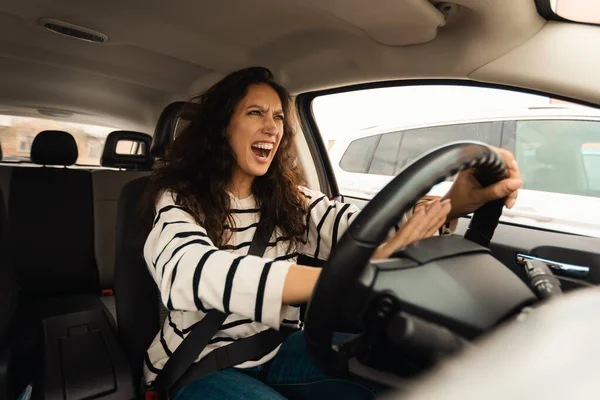 Angry female driving car shouting at somebody, emotionally reacting to accident or road jam with displeased facial expression, pressing pushing on steering wheel honking horn, windshield view