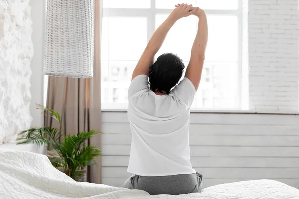 Back view of unrecognizable man in pajamas sitting on comfortable bed at home, looking through window and stretching arms, enjoying morning, feeling well-rested, copy space