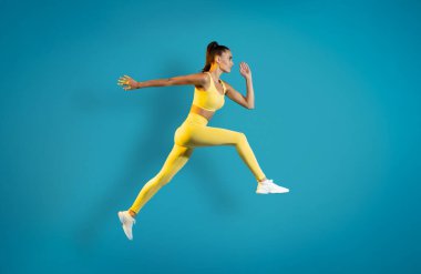 Mavi Stüdyo Arkaplanı Üzerinde Atletik Kadın Atlama Eğitiminde. Havada Sıçrayan Kadın Koşucu. Spor ve Spor Motivasyon Konsepti. Yan Görünüm, Tam Uzunluk