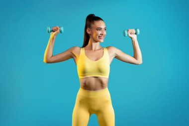 Mavi Stüdyo Arkaplanının Üzerinde Spor Poz Verirken, Sarı Spor Giysisi Giyen İyi Formalı Kadın Egzersizi Kol Kasları. Vücut Geliştirme ve Zindelik Konsepti