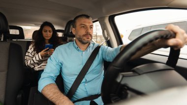 Odaklanmış erkek sürücünün portresi, arka koltukta oturan güzel bayan, cep telefonu kullanıyor, şehirde güvenli bir yolculuk yapıyor, ön cam manzaralı.