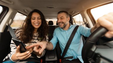 Yaz Tatili Konsepti. Lüks bir arabada oturan gülümseyen bir çiftin portresi, cep telefonunu işaret eden neşeli bir adam, yerini gösteren, dijital GPS harita uygulaması kullanan