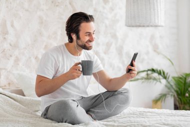 Gülümseyen yakışıklı genç adam sabah uyandıktan sonra arkadaşlarıyla görüntülü konuşma yapıyor, mutlu milenyum adamı yatakta oturuyor, akıllı telefon ve kulaklık kullanıyor, elinde bir fincan kahve tutuyor, fotokopi çekiyor.