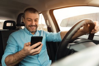 Akıllı telefon kullanan, mesajları okuyan ya da araba kullanırken e-postaları kontrol eden gülümseyen adam. Erkek sürücü cep telefonuna bakıyor, yeni cep telefonu uygulamasında daha iyi yollar arıyor, trafik sıkışıklığında.