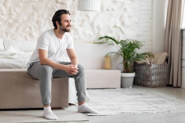 Evde pijamalı, mutlu ve sakallı genç adam pencereden bakıyor ve gülümsüyor, olumlu milenyum adamı uyandıktan sonra mutlu bir gelecek hayal ediyor, panorama.