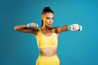 Dambıllarla Egzersiz Yapan Kararlı Fitness Kadını Sarı Spor Giysileri Giyiyor, Kollarını Kasarak Mavi Arka planda Spor Yapıyor, Stüdyo Çekimi. Spor salonu, vücut geliştirme konsepti
