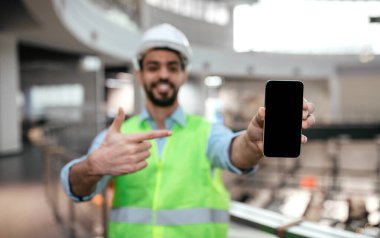 Yeni nesil İslami mühendis erkek koruyucu üniformalı, sert şapkalı, sakallı, telefonu işaret eden ve fabrika içi boş ekranlı. Reklam, uygulama ve iş, endüstri, fabrika denetimi teklifleri