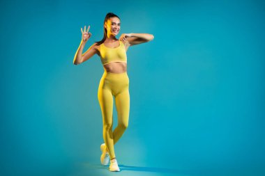 Mavi Stüdyo 'nun arkasında duran kameraya gülümseyen mutlu jest yapan kadın. Neşeli Sporcu Kadın Teklifi Onaylıyor. Sorun değil konsept. Tam Uzunluk, Boş Boşluk