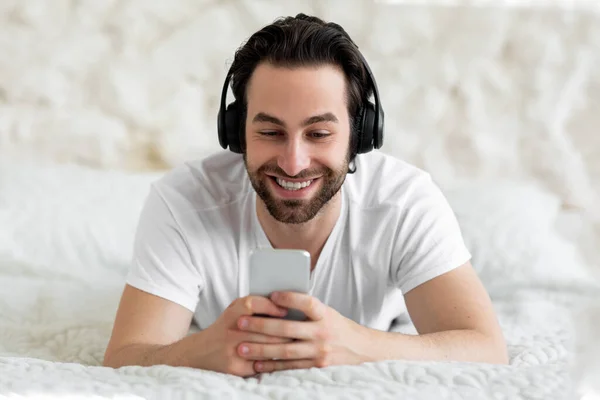 ベッドの上に寝そべっているハンサムな髭の若い男 真新しいスマートフォンとワイヤレスヘッドセットを使用して ベッドでリラックスしながらインターネット上のビデオを見て ビデオ通話と笑顔を持ち クローズアップ撮影 コピースペース — ストック写真