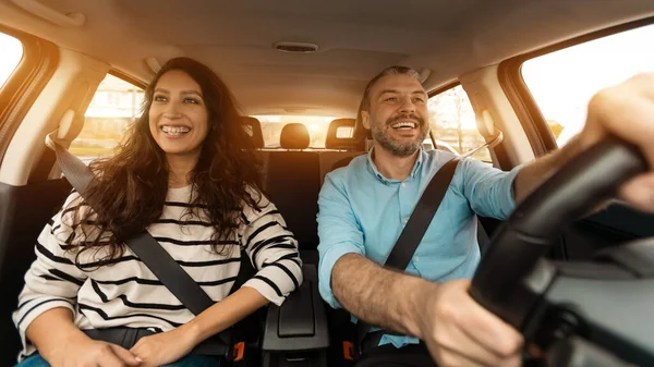 Happy man and woman going on summer vacation by car. Beautiful couple sitting in their new auto, cheerfully smiling looking at road, making test drive in the city, sun flare, windshield view