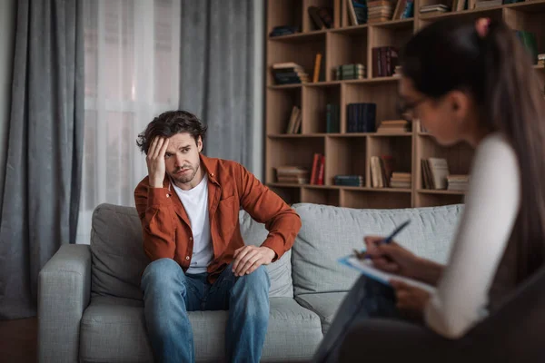 Young european female psychologist consults unhappy caucasian man in modern clinic interior. Depression, psychological problems, therapy and mental treatment, professional medical support and help