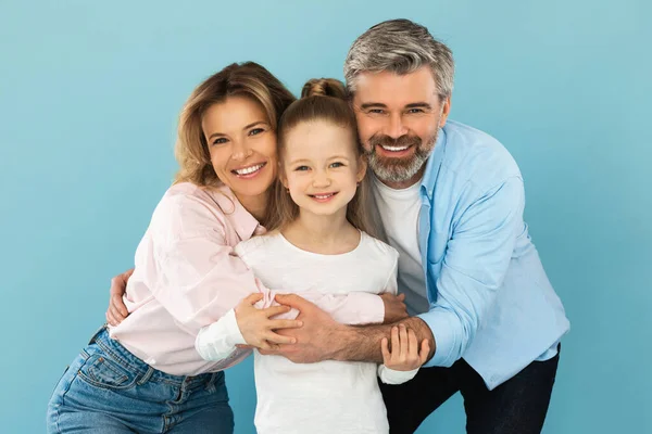 Family Happiness. Happy Middle Aged Parents Hugging Their Little Daughter Posing Standing Over Blue Studio Background, Smiling To Camera. Joy Of Parenthood Concept