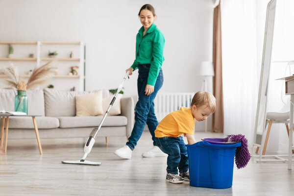Little boy helping young mother cleaning at home, child getting rag out of bucket while mom mopping floor, tidying together in living room, copy space