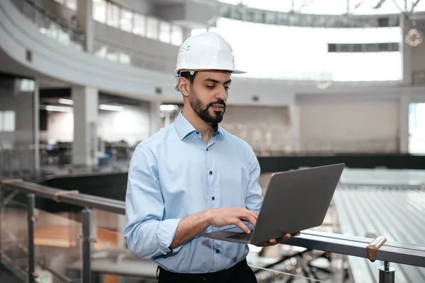 Pensive busy millennial arab guy engineer in safety helmet with beard works with computer controls factory work in plant interior. Building business, technology for industry during covid-19 outbreak