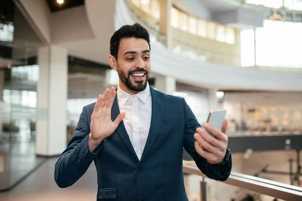 Happy millennial muslim businessman in suit with beard waving hand at smartphone webcam in office interior. Video call and technology, startup, online business, industry, finance due covid-19 outbreak