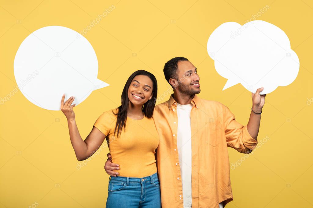 Happy young black couple holding empty speech bubbles on yellow studio background, mockup. Handsome millennial African American guy and his girlfriend posing with blank word clouds