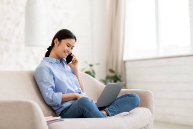 Milenyum Asyalı kadın girişimci, modern dizüstü bilgisayar kullanıyor, telefon görüşmesi yapıyor, müşterilerle görüşüyor, kucağında bilgisayarla kanepede oturuyor, akıllı telefondan konuşuyor, uzayı kopyalıyor.