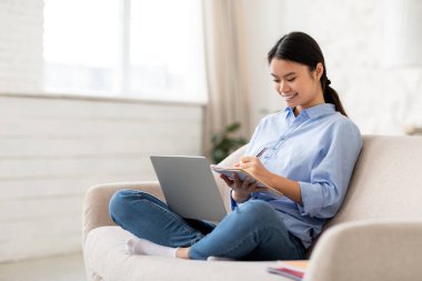 Evinden Webinar 'a katılan günlük kıyafet öğrencisi Asyalı mutlu bir milenyum kadını, koltukta oturuyor, dizüstü bilgisayar kullanıyor ve not alıyor, çevrimiçi kurslar alıyor, yan görüş, kopyalama alanı. E-eğitim kavramı