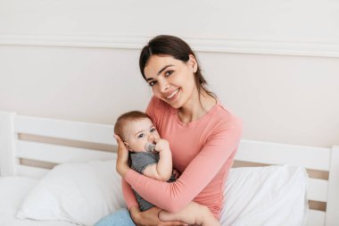 Anne ve küçük kızı. Küçük çocuğu olan genç bir anne. Çocuğu elinde tutuyor ve kameraya gülümsüyor. Evde yatakta oturuyor. Annenin yaşam tarzı, çocuk bakımı.