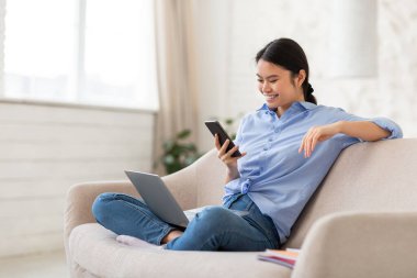 Neşeli bin yıllık Koreli serbest kadın kanepeye oturuyor, modern dizüstü bilgisayar ve yepyeni akıllı telefon kullanıyor, evden çalışıyor, müşterilerle sohbet ediyor, aletler kullanıyor, kopyalama alanı kullanıyor, yan görüş kullanıyor.