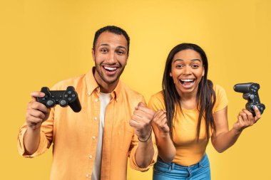 Boş zaman eğlence konsepti. Heyecanlı genç siyah çift video oyunları oynuyor, joystickleri tutuyor ve kullanıyorlar, oyun kazanıyorlar, sarı stüdyo arka planında EVET işareti yapıyorlar.