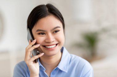 Yakın plan portresi, mavi gömlekli, cep telefonuyla erkek arkadaşıyla konuşan, fotokopi odasına bakan, arka planı bulanık, panorama olan çekici Asyalı bir kadın. İletişim konsepti