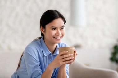 Evdeki kanepede oturmuş, bitki çayı ya da kahve içen, fotokopi odasına bakıp gülümseyen ve eğlenen güzel mavi tişörtlü Asyalı kadının portresi.