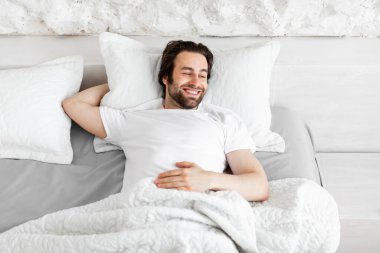 Neşeli, yakışıklı, sakallı, milenyum sakallı genç adam yatakta tek başına yatakta uzanıyor, beyaz pijama giyiyor, fotokopi odasına bakıyor ve gülümsüyor, yukarıdan bakıyor.