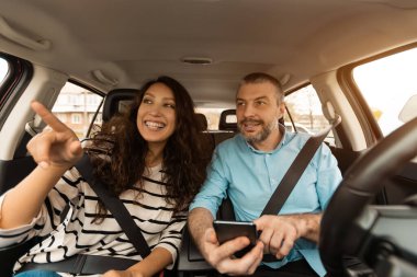 Yol Yolculuğu Konsepti. Arabada oturan gülümseyen bir çiftin portresi, araba kullanan ve cep telefonu kullanan bir erkek, yolu gösteren bir kadın, dijital harita uygulaması kullanan, ön cam manzaralı bir kadın.