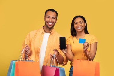 Havalı genç siyah çift ellerinde alışveriş torbaları ve kredi kartı tutarken sarı ekranlı akıllı telefon, yeni online alışveriş uygulaması tasarımı için model.