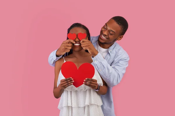 愛の概念によって盲目にされる 愛情深いアフリカ系アメリカ人のカップルは レッドペーパーハートを保持します 彼のガールフレンドの目をカバー笑顔男 ピンクのスタジオの背景に隔離された楽しさを持っているロマンチックな男と女 — ストック写真