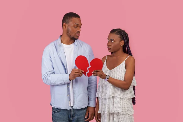 爱的终结概念 不快乐的非洲裔美国夫妇抱着两片破碎的红心 孤零零地站在粉红色的工作室背景墙上 正在经历离婚或破裂的年轻家庭感到不安 — 图库照片