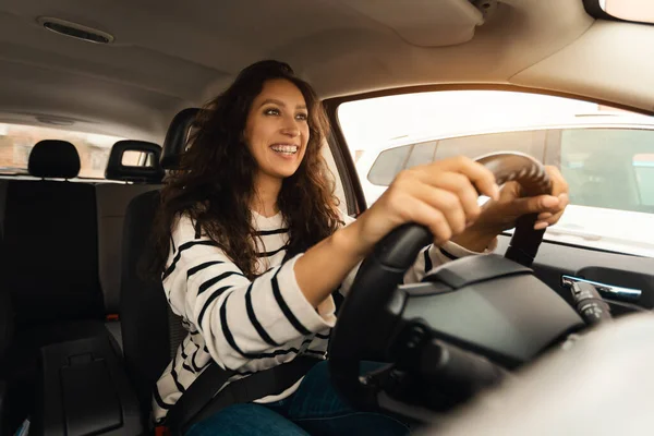 Confident Professional Driver. Portrait of happy young woman sitting in luxury car on drivers seat. Positive black lady riding in the city holding hands on steering wheel, looking at road