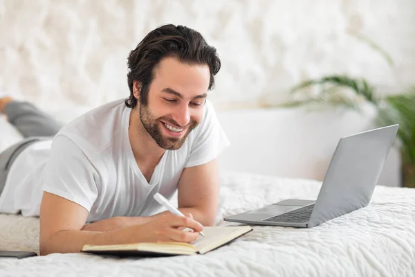 Smiling bearded young man in pajamas laying on bed, using laptop and taking notes, unemployed guy looking for job on Internet, home interior, side view, copy space. Job search website