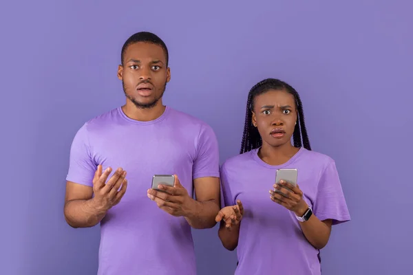 コンテンツの設定または妨害 混乱した千年のアフリカ系アメリカ人のカップルの肖像画彼らの携帯電話を保持し 肩をカメラを見て 紫色の壁に隔離された立っている — ストック写真