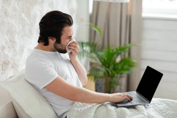 Sick young man in pajamas laying in bed, sneezing runny nose and using modern laptop with black empty screen, looking for COVID-19 symptoms on Internet, mockup, side view, copy space