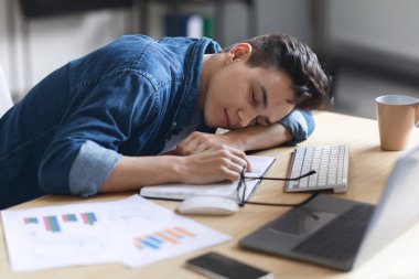 Aşırı çalışma kavramı. Yorgun erkek çalışan ofiste uyuyor, yorgun milenyum adamı iş yerinde kestiriyor, masaya yaslanıyor, elleri üzerinde yatıyor, açık not defterine uzanıyor, yakın çekim.