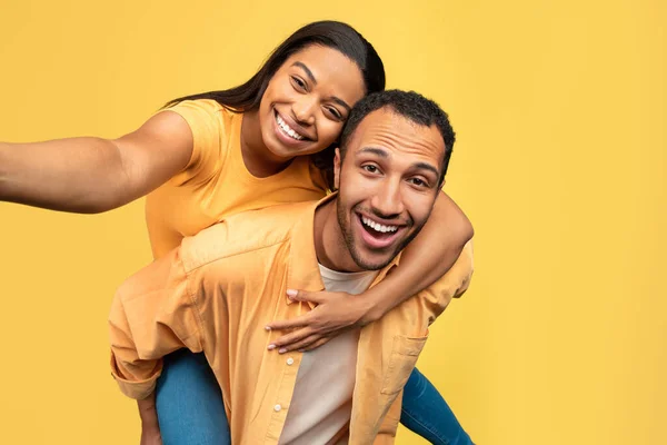 很酷的年轻黑人家伙让他的女朋友搭便车 使自拍的黄色工作室背景 有爱心的非洲裔美国人夫妇一边拍照一边玩乐 — 图库照片