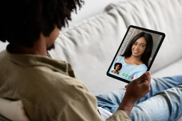 Mobile application for telecommunication concept. Millennial african american man using modern digital tablet, sitting on sofa at home and calling his beautiful girlfriend or wife, over shoulder shot