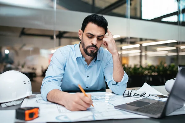 Sad serious pensive attractive millennial arab man engineer with beard works with project drawings at workplace with hard hat and pc in office interior. Brainstorm, business, industry, construction