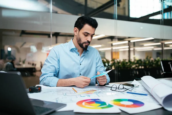 Busy serious attractive millennial arab guy designer with beard works with color palettes at workplace with computer in office interior. Brainstorming, business, industry project, work due covid-19