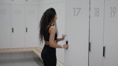 Başarılı fitness egzersizi. Genç, aktif bir bayan, dijital bilezik anahtarıyla dolabı açıyor ve ceketini alıyor. Spor eğitiminden sonra soyunma odasında giyiniyor. Ağır çekimde, boş alanda.