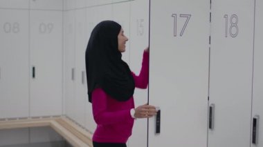 Başörtülü genç aktif Müslüman kadın akıllı telefonu dolaptan alıyor, mesajları kontrol ediyor ve spor salonundaki soyunma odasında anahtarlıkla kapatıyor, ağır çekimde.